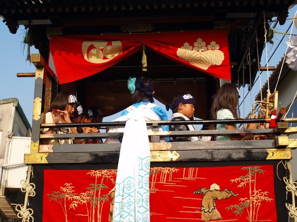 神輿の中で笛を演奏している女の子、ハチマキをしている男の子が乗っている写真